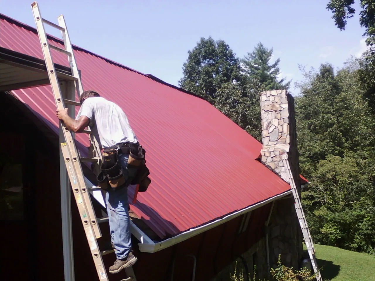 Roofer on a ladder beside a red metal roof and stone chimney — Soffit & Fascia Repair by Roofing Brothers in Unicoi